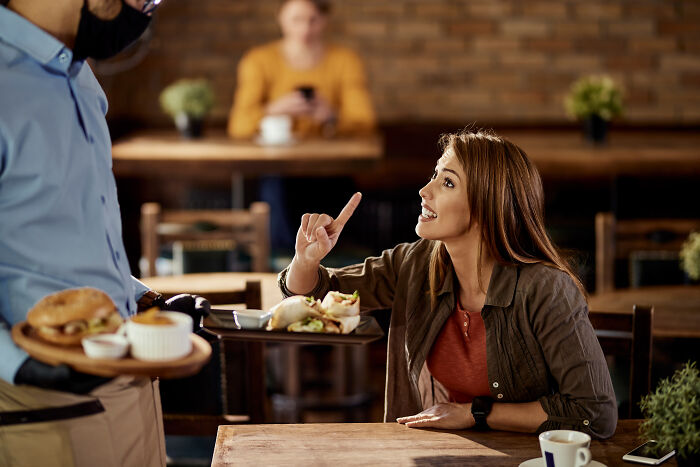 A young woman in a café talking to a waiter, showing signs of having grew up sheltered in her behavior.