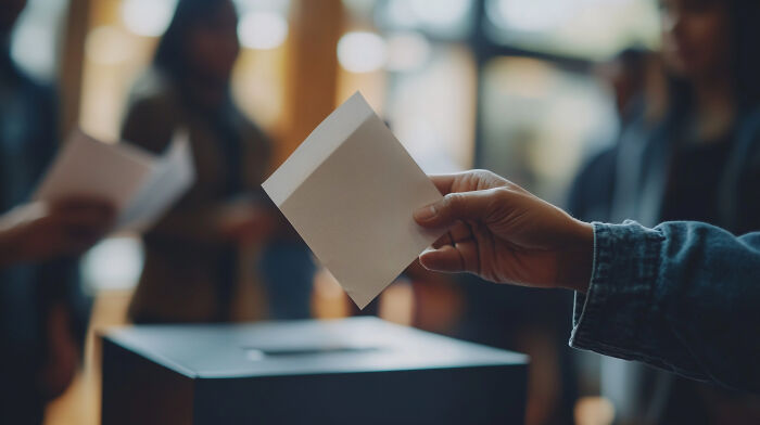 Person holding paper ballot above a box, with blurred figures in the background, illustrating signs grew up sheltered concept.