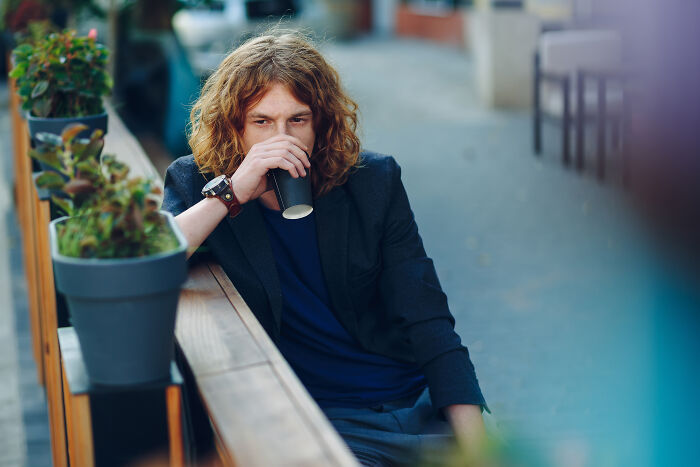 Young man sitting alone outdoors, looking thoughtful while drinking coffee, illustrating signs grew up sheltered.