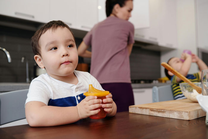 Young child with sippy cup sitting at the table while mother busily attends to kitchen tasks in background.