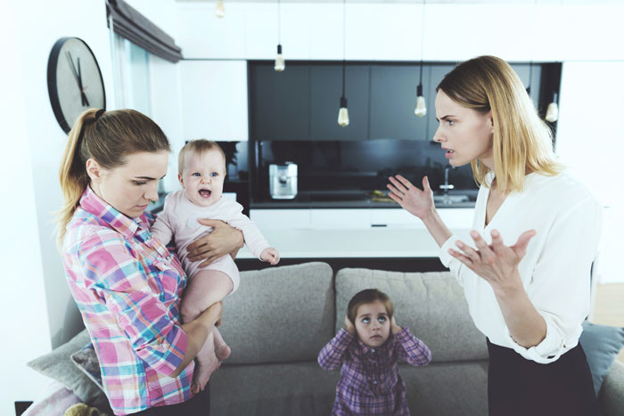 Young woman holds baby while facing sister-in-law setting pregnancy husband boundaries in modern living room. - 29