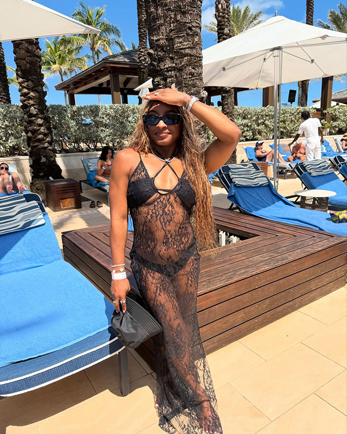 Simone Biles in a black lace swimsuit cover-up, wearing sunglasses and a bracelet, posing poolside during a sunny vacation.