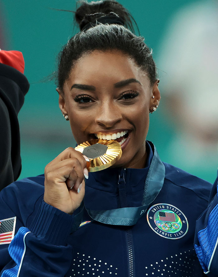 Simone Biles smiling and biting a gold medal while wearing Team USA jacket, sparking plastic surgery rumors