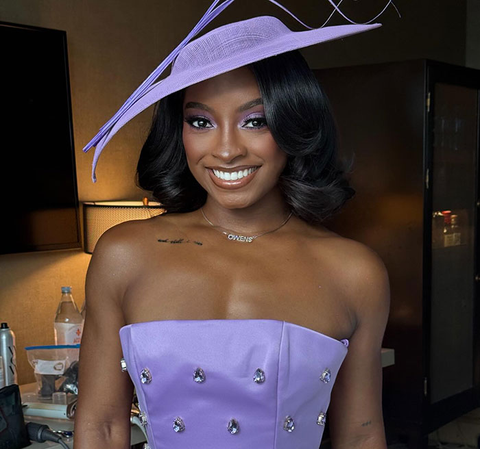 Simone Biles wearing a lavender strapless top and matching hat, sparking plastic surgery speculation with bikini pics.