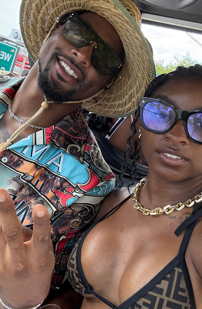 Simone Biles wearing sunglasses and bikini while posing with a man in a straw hat and patterned shirt outdoors.