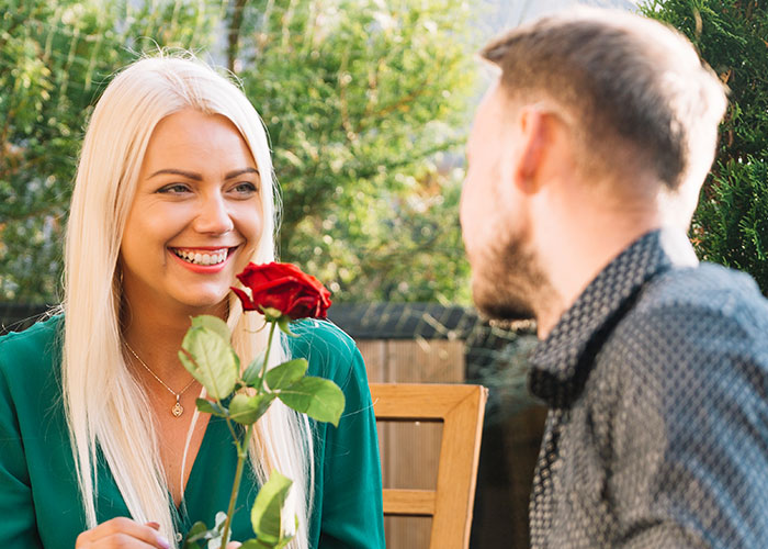 A single woman smiling while holding a red rose, avoiding men in an outdoor dating setting.