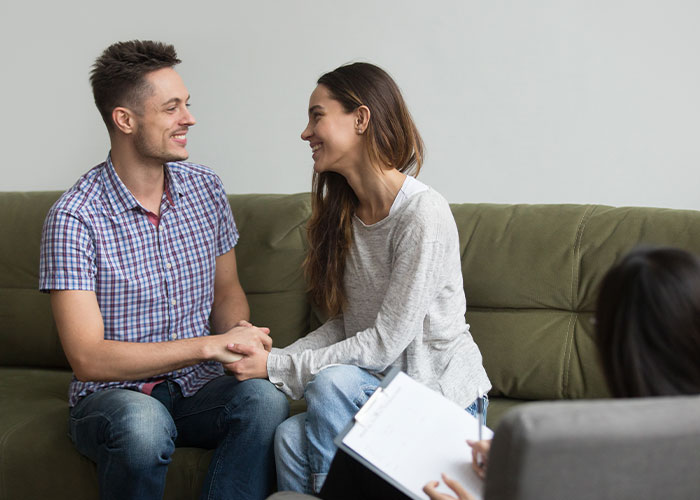 Couple sitting on a couch smiling and holding hands during a relationship counseling session with a therapist.