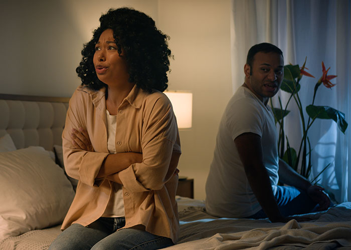 Single woman avoiding a man sitting on bed, showing frustration and emotional distance in a dimly lit bedroom.