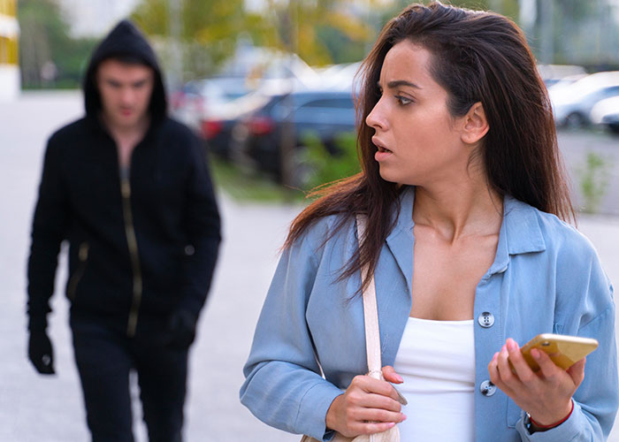 Young single woman looking worried while holding phone as a man in a hoodie walks behind her outdoors, avoiding men.