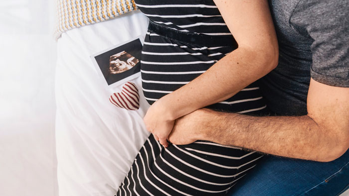 Pregnant woman in striped dress embraced by partner with ultrasound photo on bed, symbolizing baby and conception struggles.
