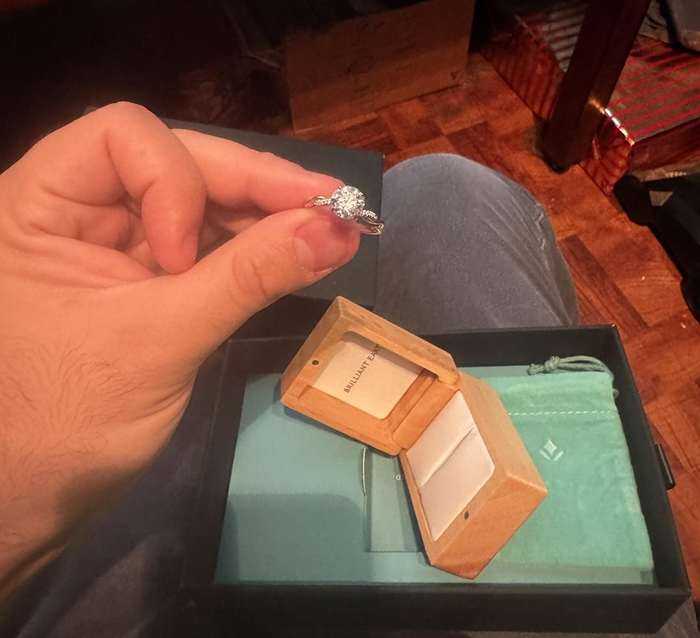 Hand holding a small propose ring over an open wooden ring box, with a gift bag and presents in the background. Hand holding a small propose ring over an open wooden ring box, with a gift bag and presents in the background.