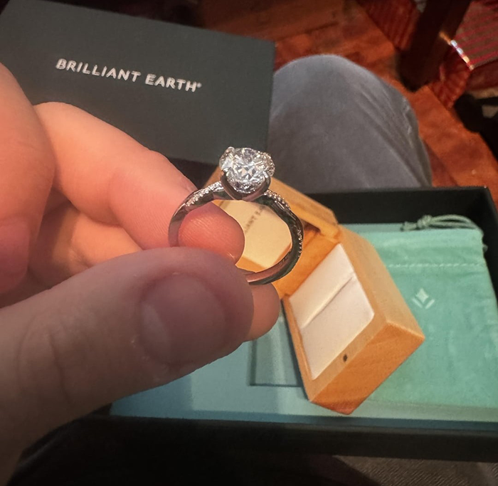 Hand holding a small girlfriend proposal ring with a diamond, next to an open wooden ring box and Brilliant Earth packaging. Hand holding a small girlfriend proposal ring with a diamond, next to an open wooden ring box and Brilliant Earth packaging.