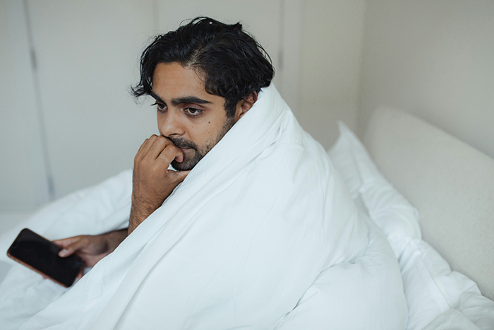 Man wrapped in a white blanket sitting on bed looking pensive while holding a phone, reflecting on small propose ring choice Man wrapped in a white blanket sitting on bed looking pensive while holding a phone, reflecting on small propose ring choice