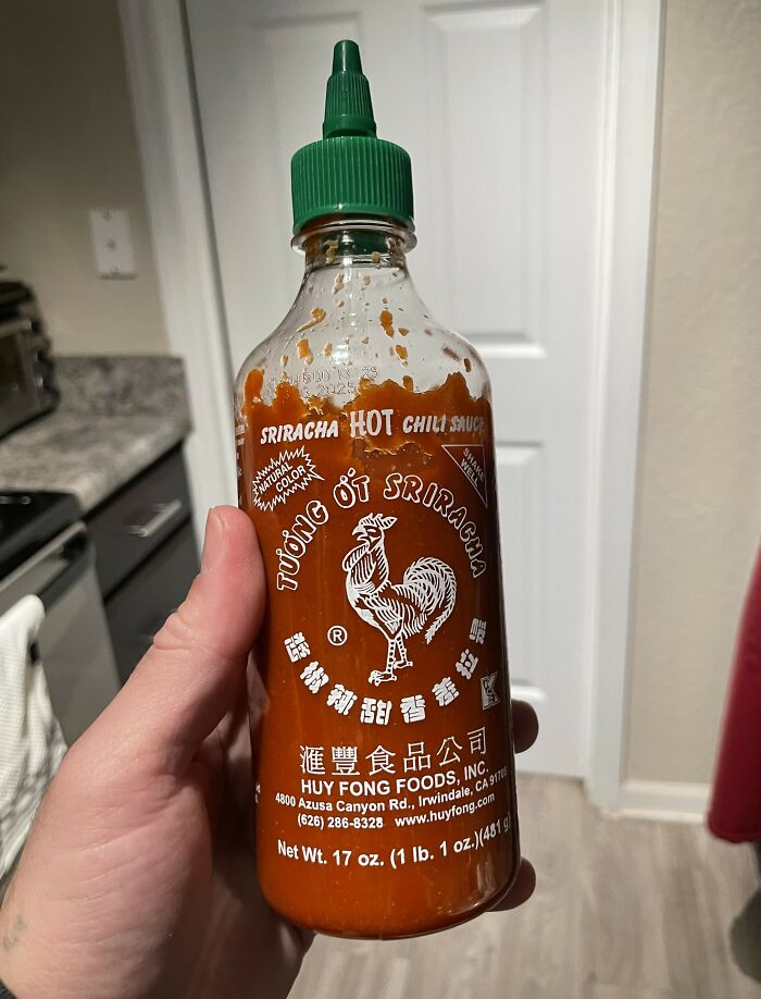 Hand holding a nearly empty Sriracha hot chili sauce bottle in a kitchen setting, from poverty to super wealthy.