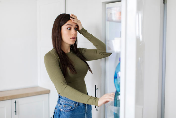 Woman looking distressed opening fridge, reacting to ruined custom baby shower cake in shared fridge at home.