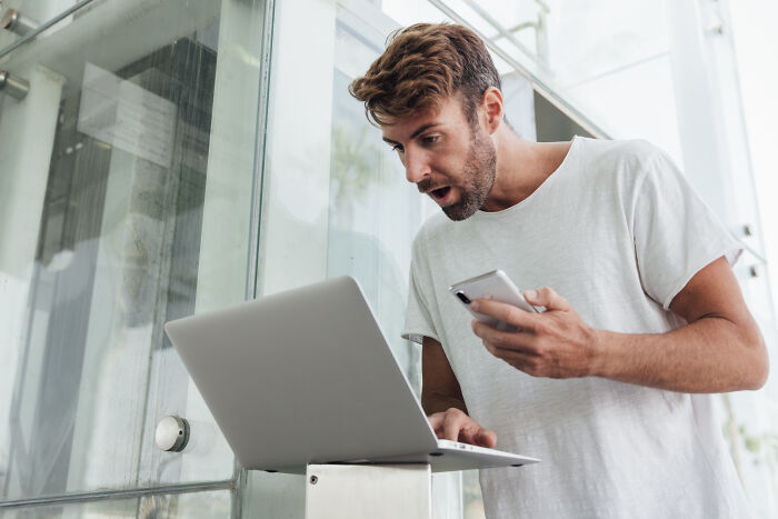 Man in white shirt shocked looking at laptop and phone, illustrating mortifying stories of sending messages to wrong person. - 3