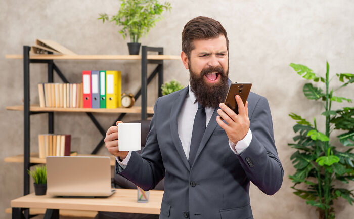 Bearded man in suit looking shocked at phone while holding coffee mug, illustrating sending something to wrong person stories. - 13
