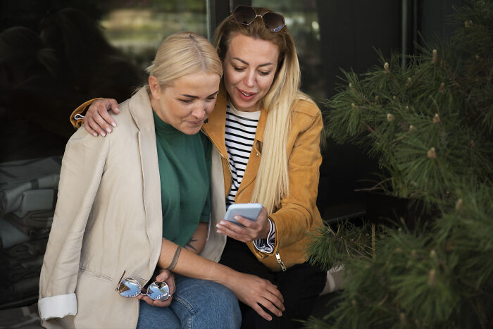 Two women looking at a phone together, reacting with surprise, illustrating mortifying stories of sending to the wrong person. - 4