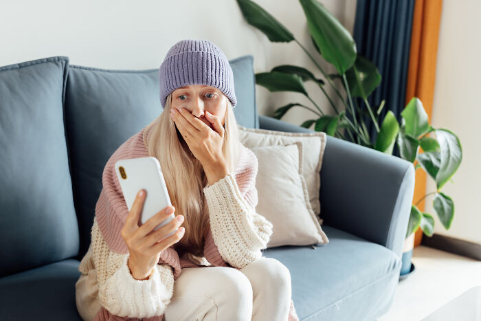 Woman looking shocked at phone, sitting on a couch, depicting mortifying stories of sending messages to wrong person. - 7