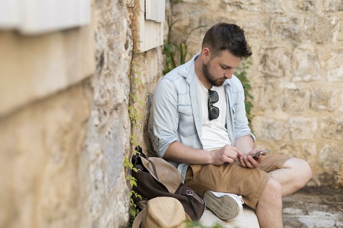 Young man sitting against a stone wall, looking at his phone, reflecting on mortifying stories of sending to wrong person. - 28