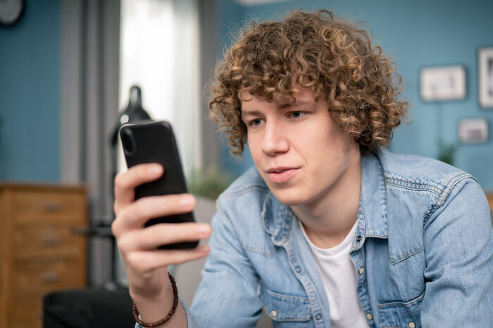 Young man with curly hair in a denim jacket looking at his phone, illustrating a mortifying story of sending to the wrong person. - 14