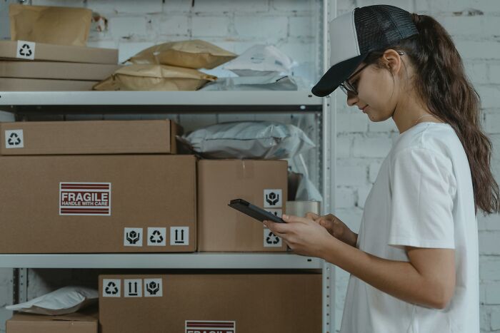 Woman wearing cap with glasses using phone in a shipping storage area filled with fragile packages, showing sending something wrong person. - 31
