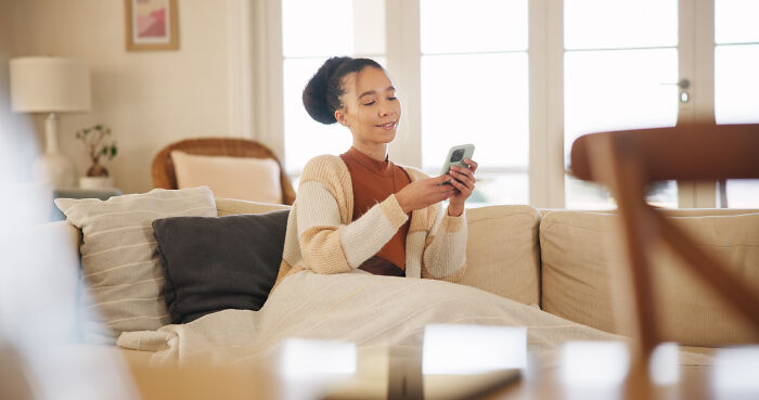 Young woman sitting on couch, smiling while checking phone, illustrating stories of sending messages to wrong person. - 30