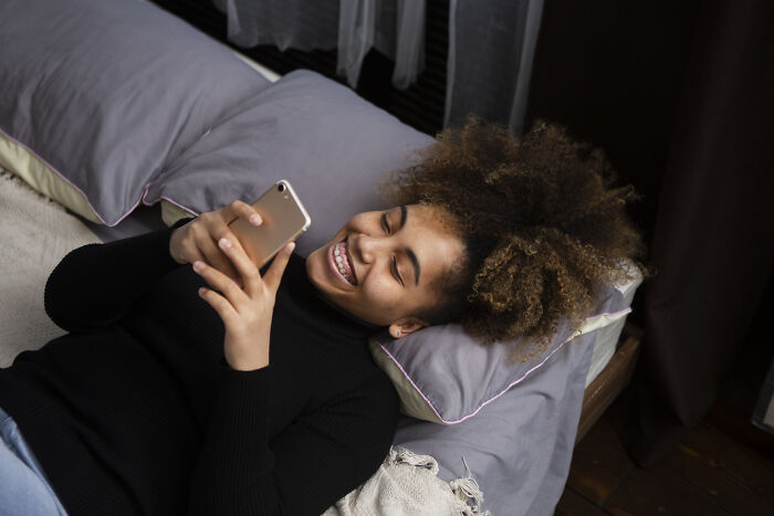 Young woman lying on bed, smiling while using smartphone, illustrating relatable stories of sending messages to the wrong person. - 5