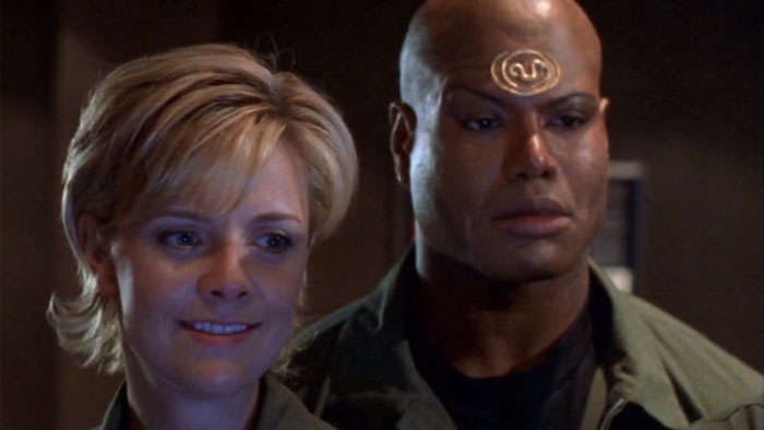 Two Stargate characters in military uniforms, one smiling and the other serious, in a dimly lit room.