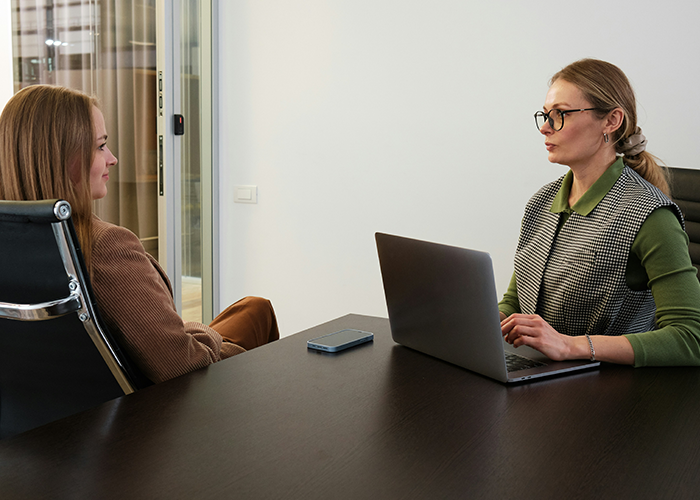 Boss struggling with technology confronts coworker in office, sitting across a desk with laptop and smartphone nearby.