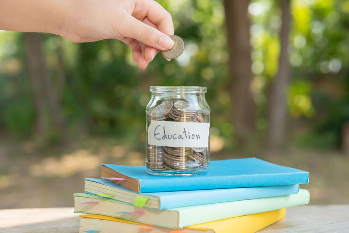 Hand placing a coin into an education jar on a stack of books, symbolizing saving and stealing daughters trust fund.