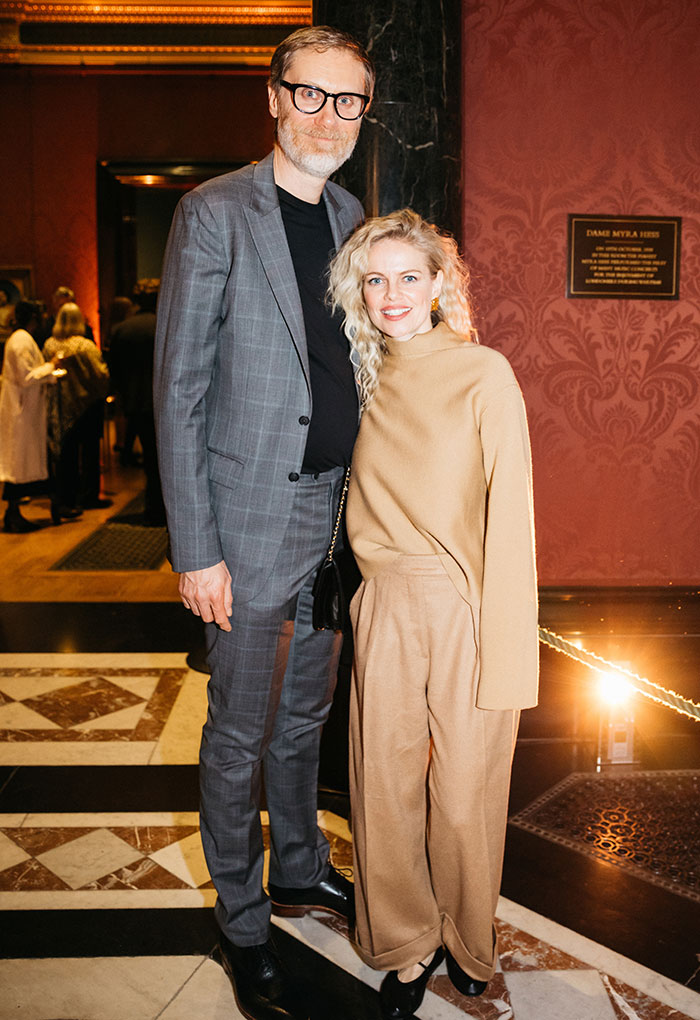 Tall male star wearing glasses and gray suit standing next to a woman in beige outfit showcasing super tall celebrity height difference.