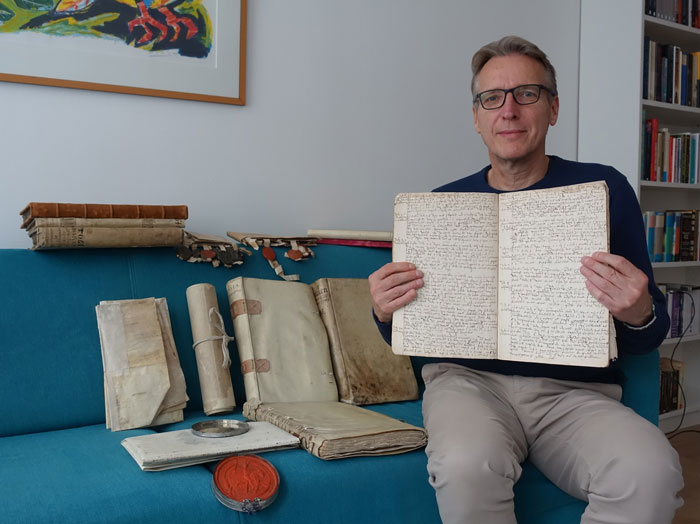Man sitting on a couch holding a large historic document with other old manuscripts and books around him. - 1