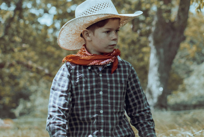 Young boy wearing a cowboy hat and bandana outdoors, evoking childhood stories that were really messed up.