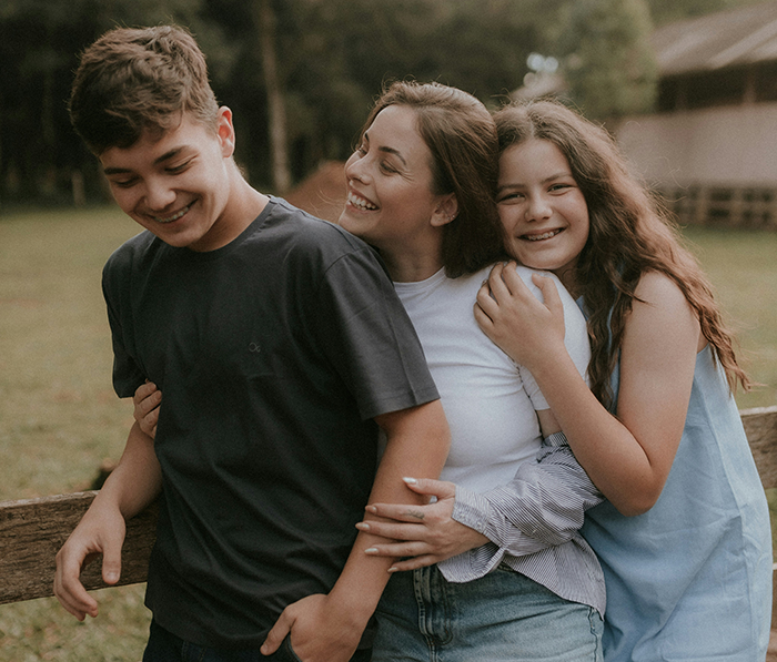 Student working mom happily hugging her two children outdoors in a park setting with natural green background.