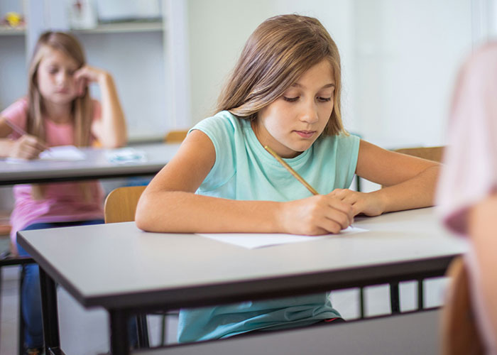 Young girl focused on writing at school desk while other students work in the background, reflecting school punishments concerns. - 11