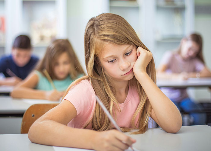 A schoolgirl looking upset while writing at her desk, with other students focused on their work in the classroom. - 29