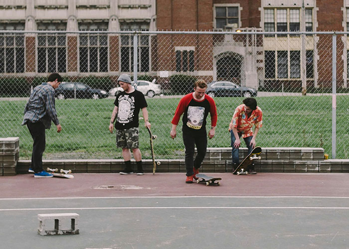 Four teenagers skateboarding and hanging out near a schoolyard fence, reflecting on school punishments. - 2