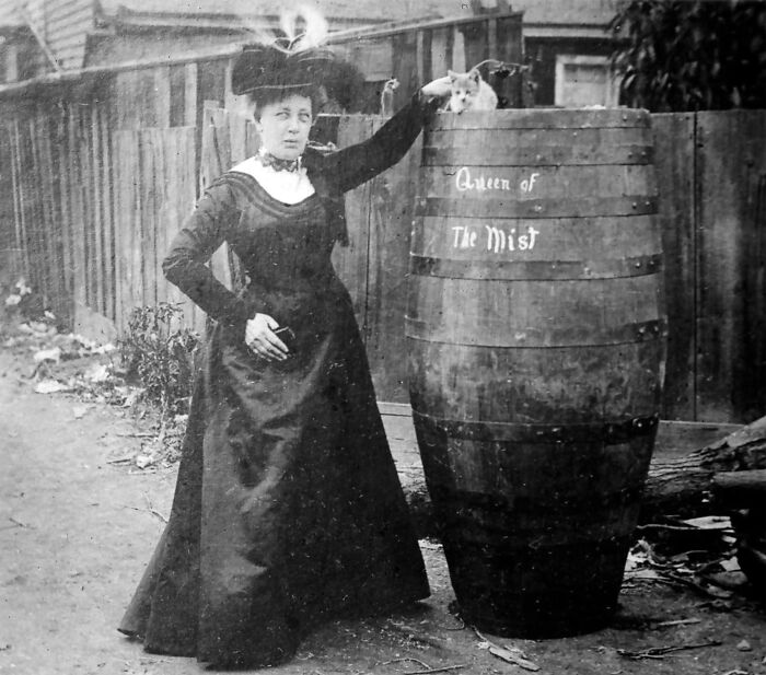 Woman in Victorian dress posing with cat on barrel labeled Queen of The Mist in unique old photo outdoors.