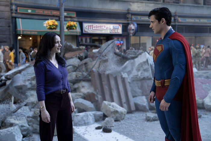 Superman in blue suit and red cape talks to a woman amid rubble, highlighting woke Superman reboot in early reviews.