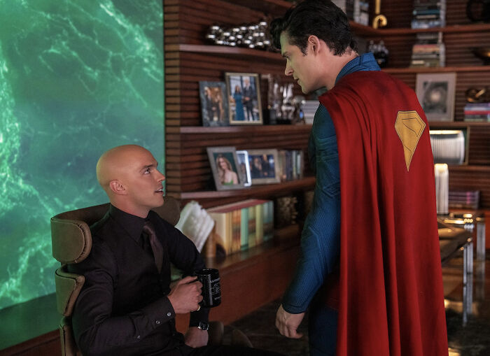 Superman reboot scene with man in black suit holding mug facing superhero in blue suit and red cape indoors.