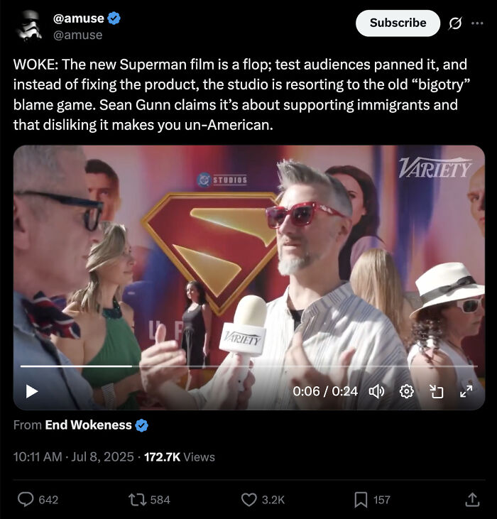 Man with gray hair and red sunglasses interviewed at Superman movie premiere with logo backdrop, discussing woke Superman reboot flop.