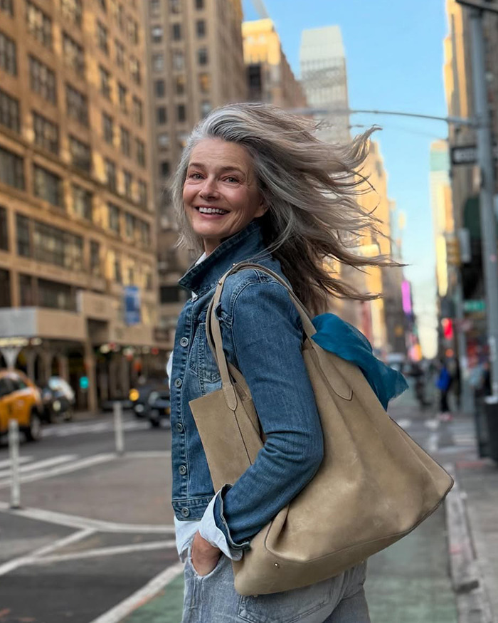 Supermodel 60 smiling confidently on city street wearing denim jacket and carrying beige tote bag with windblown hair. - 1