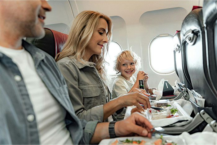 Woman sneakily swaps airplane seats to avoid babysitting little sister during family trip paid by dad, smiling while eating.