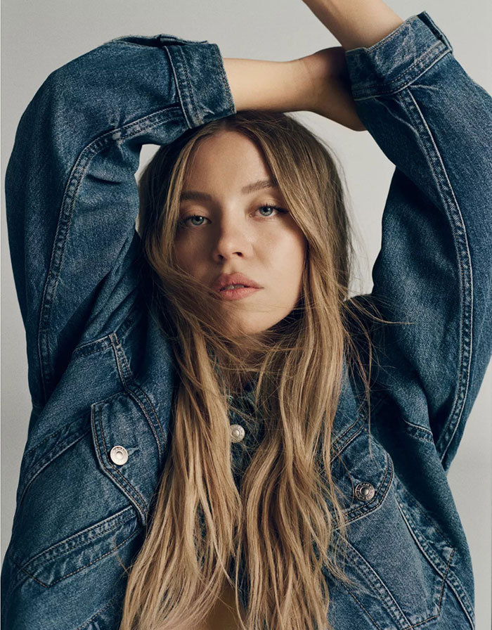 Young woman in a denim jacket posing with arms raised, related to Sydney Sweeney woke scandal and ethnic cleansing controversy. Young woman in a denim jacket posing with arms raised, related to Sydney Sweeney woke scandal and ethnic cleansing controversy.