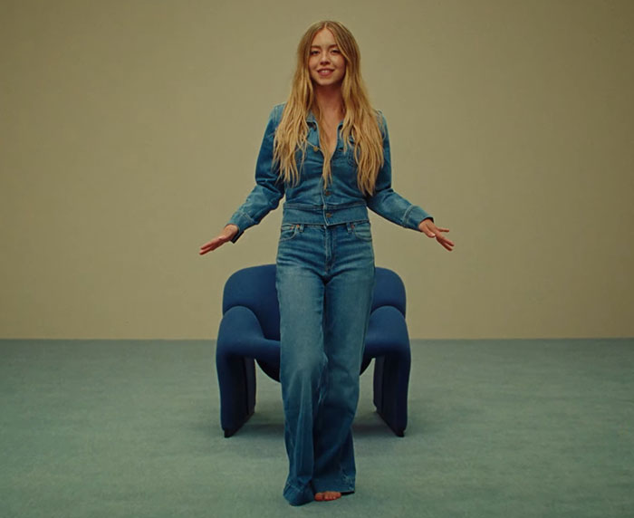 Sydney Sweeney standing barefoot in denim outfit in front of modern blue chair, sparking woke scandal and ethnic cleansing debate. Sydney Sweeney standing barefoot in denim outfit in front of modern blue chair, sparking woke scandal and ethnic cleansing debate.