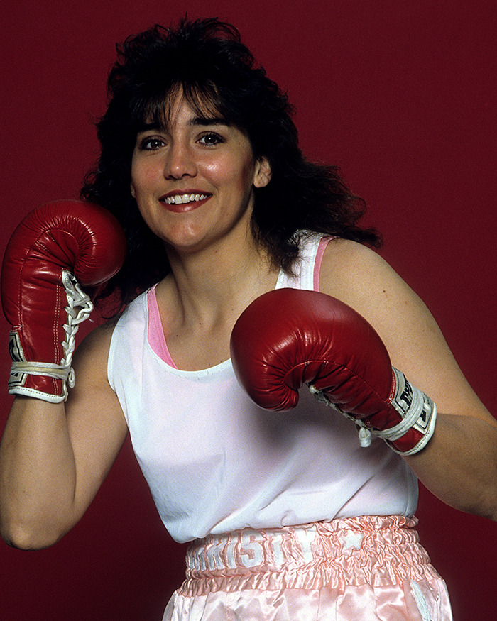 Female boxer wearing red boxing gloves and pink shorts posing against maroon background for Sydney Sweeney body transformation role. Female boxer wearing red boxing gloves and pink shorts posing against maroon background for Sydney Sweeney body transformation role.
