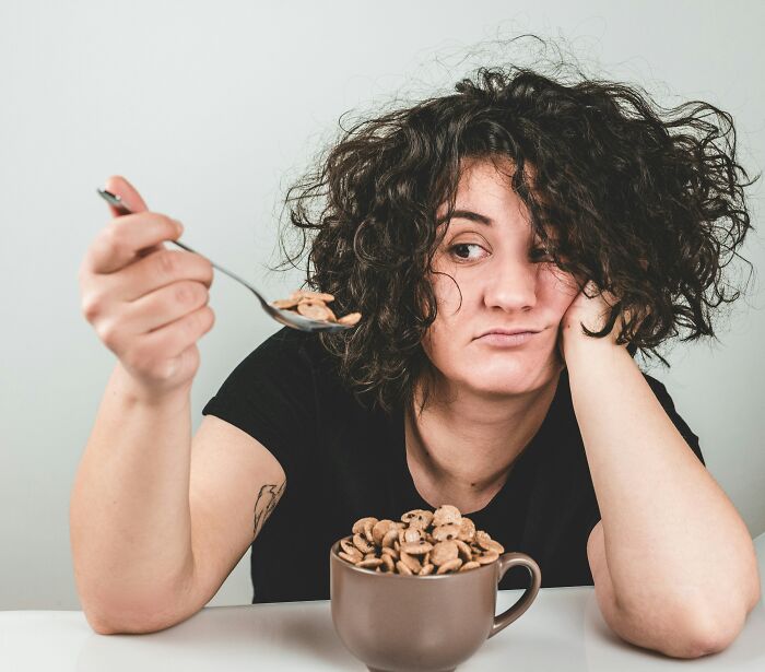 Young woman with messy hair looking unenthusiastic while holding a spoon over a cup of cereal, reflecting parent harmless lies trauma. - 8
