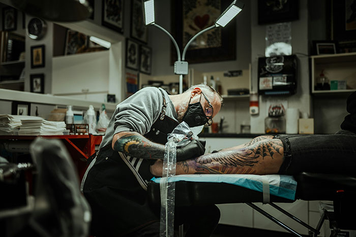 Tattoo artist wearing a mask carefully working on a large leg tattoo in a detailed tattoo studio environment.