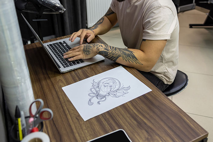 Tattoo artist with sleeve tattoos working on laptop near custom design sketch in a creative studio space.